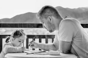 Black and white picture of a father and young daughter drawing on a porch overlooking some mountains.