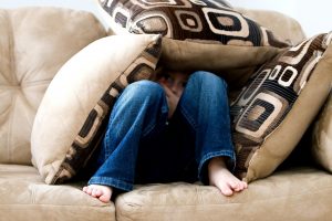 A boy in a blue denim pants is sitting on a couch, hiding under three pillows because he's struggling with adoption